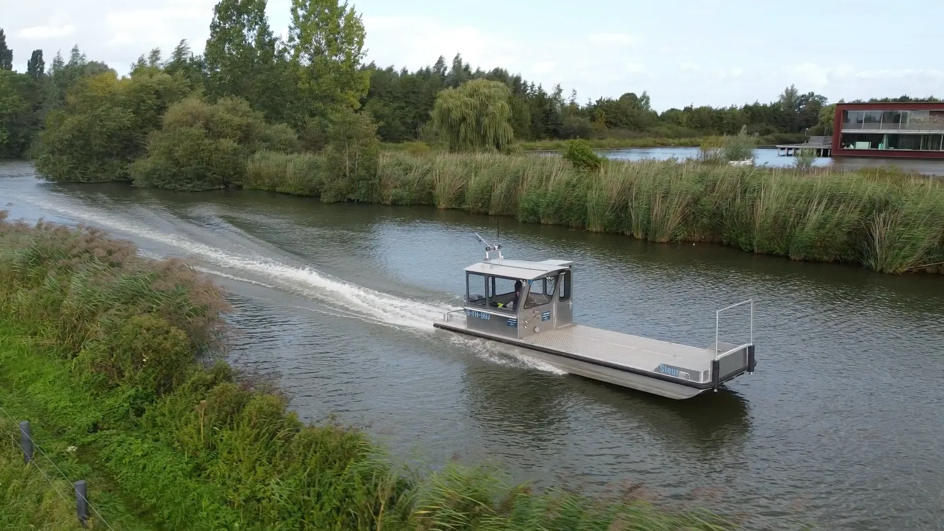 Dronebeeld van werkboot Steur die met kielzog langs een rietoever vaart, met bomen en water op de achtergrond