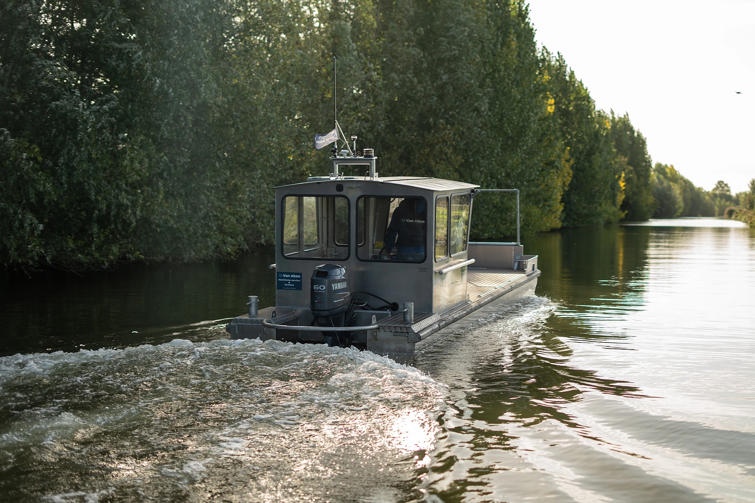 Steur vaart weg langs een bomenrij, met kielzog achter de motor